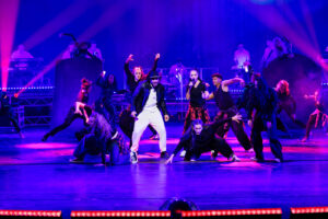 Tanzgruppe auf Bühne in bunten Lichtern performt lebhaft.