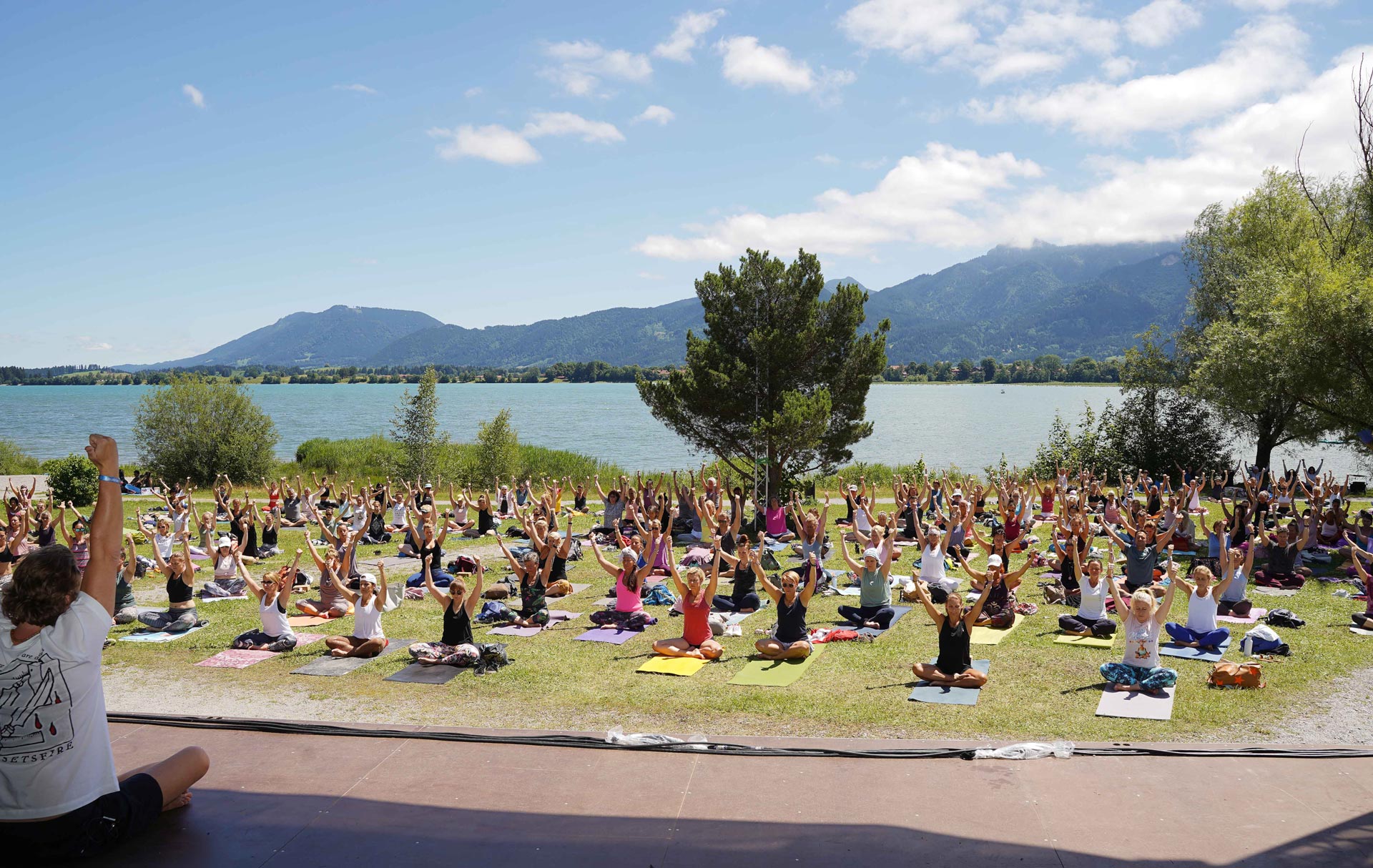 Yoga-Gruppe am See mit Bergen im Hintergrund.